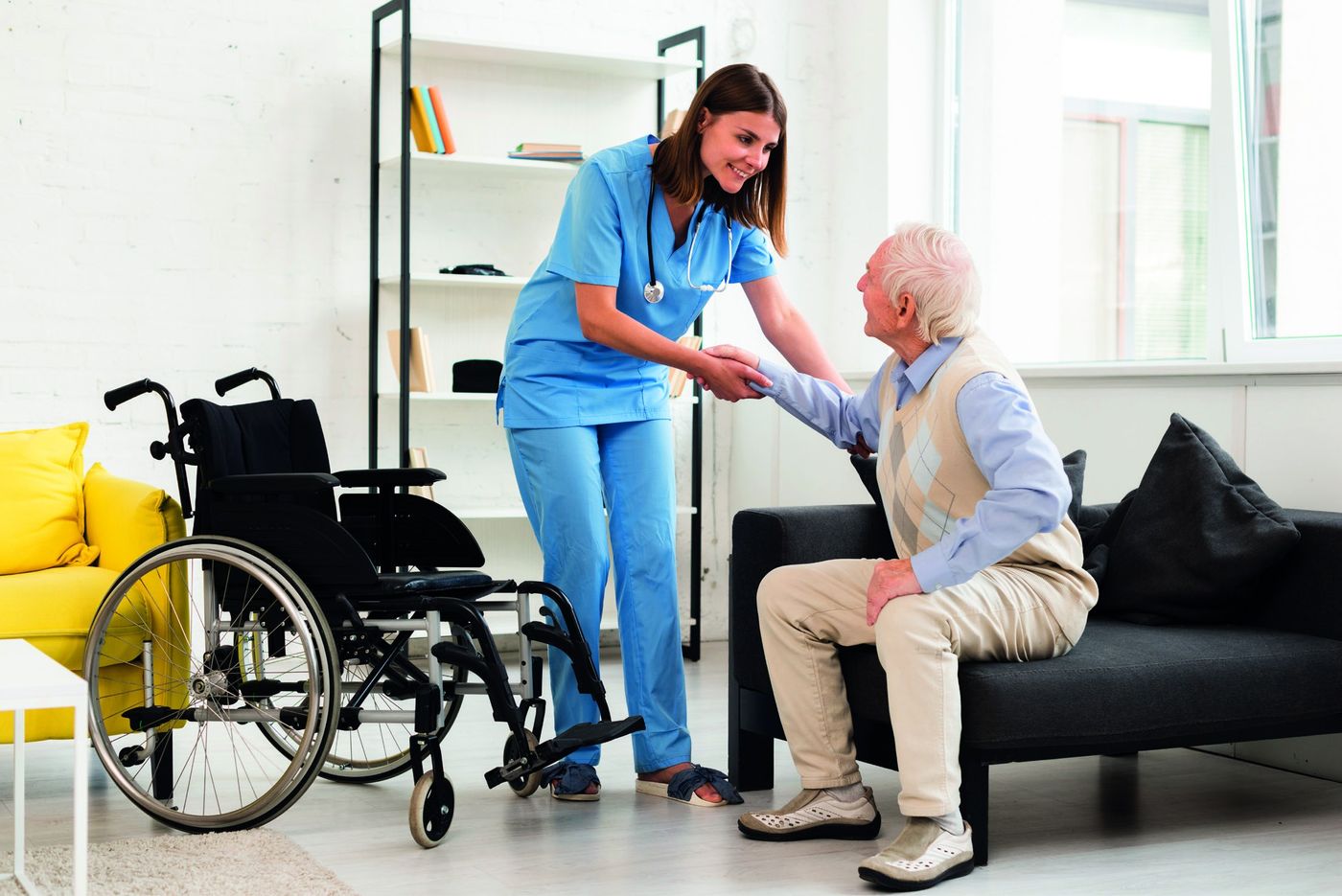 A smiling nurse helps an elderly man rise from his chair at home