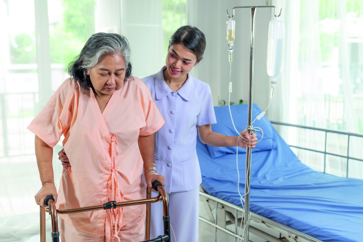 A caregiver supports an older woman walking with a walker beside a hospital bed